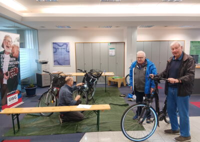 Mehrere Männer nehmen an einem Fahrrad-Workshop teil. Einer kniet und repariert ein Rad, zwei andere stehen mit ihren Fahrrädern