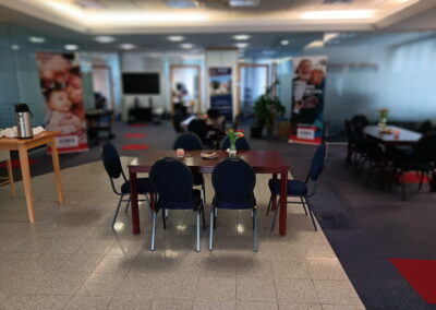 Besprechungsraum mit Holztisch, Stühlen, Blumen und Snacks im Vordergrund. Im Hintergrund weitere Tische und unscharfe Poster